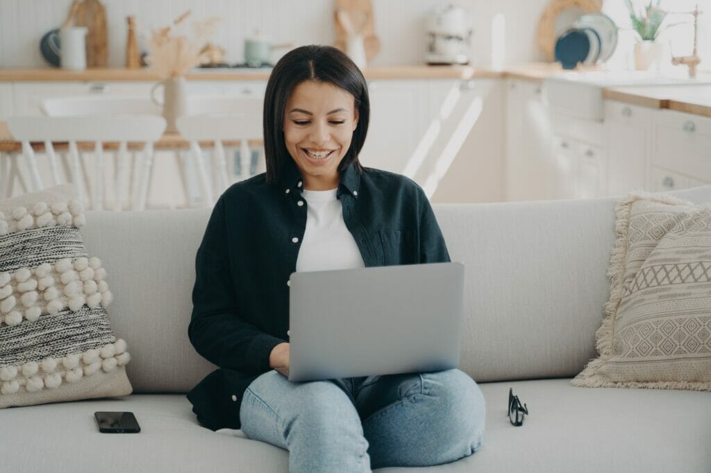 Smiling female shopping on the internet at laptop sitting on couch at home. Ecommerce platform malaysia, online store